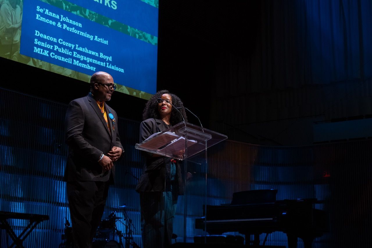 Two people, a man and a woman, stand on a stage behind a clear podium with papers on it. The man wears a dark suit with glasses, and the woman, wearing glasses and a dark outfit, looks at him. A large screen in the background displays text with names and titles. The stage is dimly lit with blue lighting.